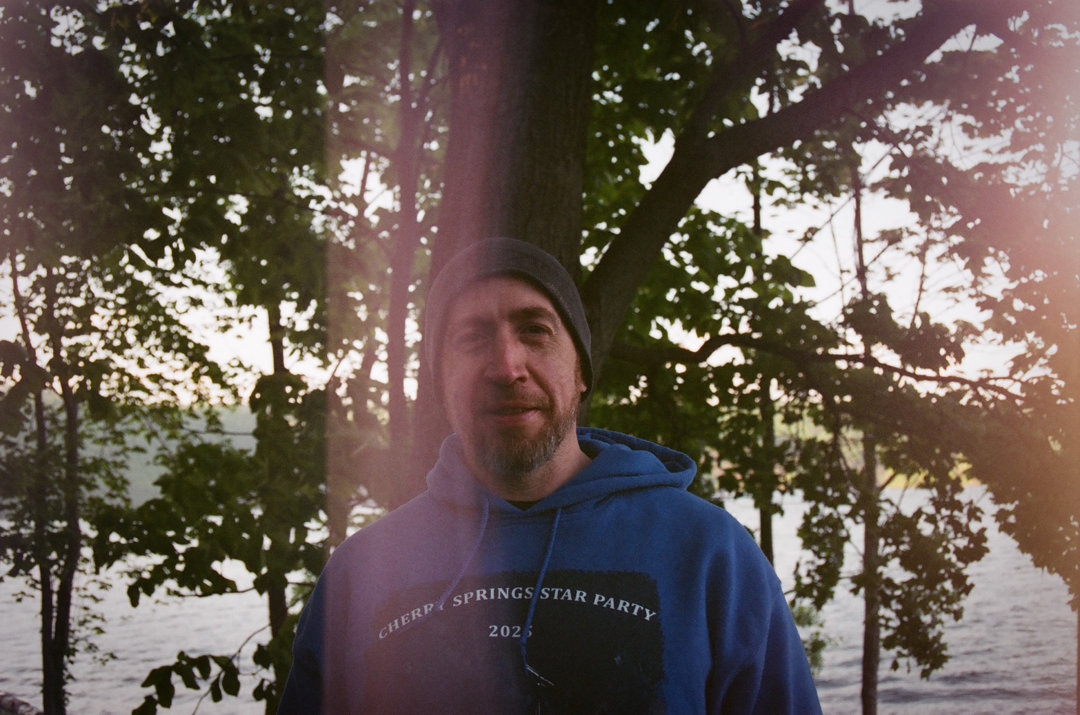 Bill McGeeney in a gray beanie and blue Cherry Springs Star Party hoodie, standing among trees beside a lake at dusk